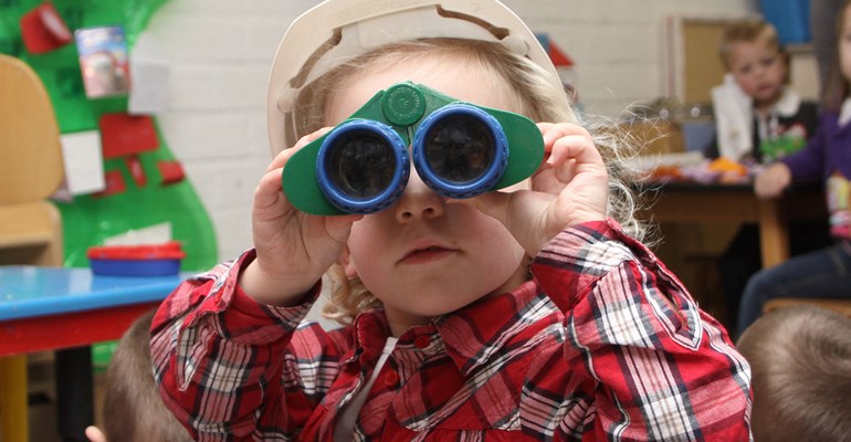 Kind kijkt op de kinderopvang door verrekijker naar de camera.